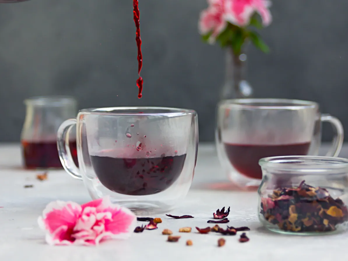 tasse transparente avec décoction d'hibiscus versée dedans et au première plan des fleurs d'hibiscus