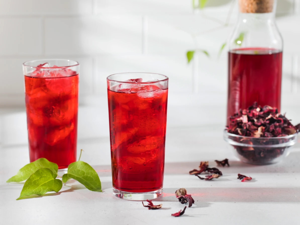 Infusion à froid d'hibiscus avec 2 verres de bissap ainsi qu'une bouteille posés sur une table avec des fleurs séchées dans un bol