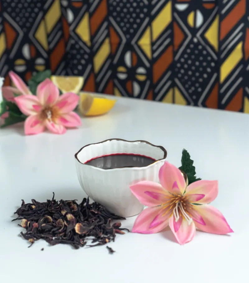 fleurs d'hibiscus sur une table avec une tasse de bissap, en arrière plan tissu bogolan wax