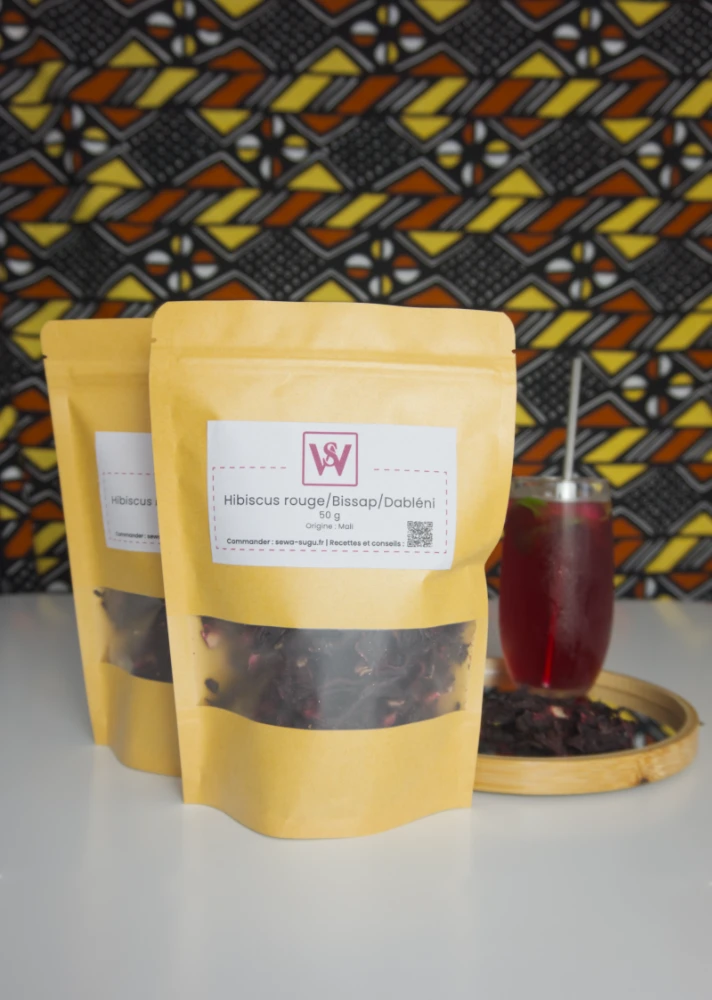 deux paquets krafts de fleurs d'hibiscus séchées sur une table blanche avec du wax en fond et un verre d'hibiscus sur le côté